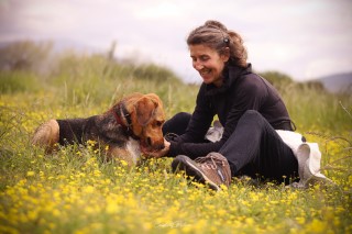 Une femme riant avec bonheur tout en jouant avec son chien dans un champ de fleurs jaunes, capturant un instant de complicité et de détente dans la nature.